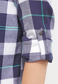 Navy and white plaid shirt with green accents. Features herringbone pattern, rolled sleeves, and a decorative button on the cuff.