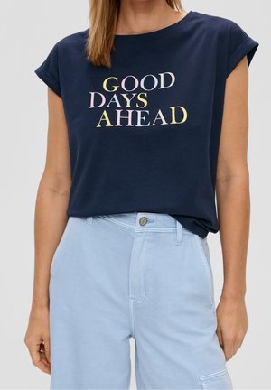 Kvinde iført en marineblå skjorte med opkærmede ærmer og pastelteksten "GOOD DAYS AHEAD", parret med lyseblå højtaljede bukser.