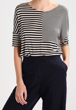 Camiseta a rayas negras y crema con mangas cortas, escote redondo y un diseño dividido que presenta anchos de raya variados.