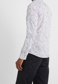 Biała koszula z długim rękawem z czarnym abstrakcyjnym wzorem linii, z kołnierzykiem zapinanym na guziki i guzikami przy mankietach, wykonana z lekkiego materiału, o dopasowanym kroju.