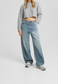 Vrouw in een cropped grijze kabelgebreide sweater, losse lichtblauwe jeans en grijs-witte sneakers, met een hand in de zak.