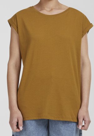 Persona con una camisa de manga corta color mostaza con los puños remangados y vaqueros de mezclilla azul claro, de pie contra un fondo liso.