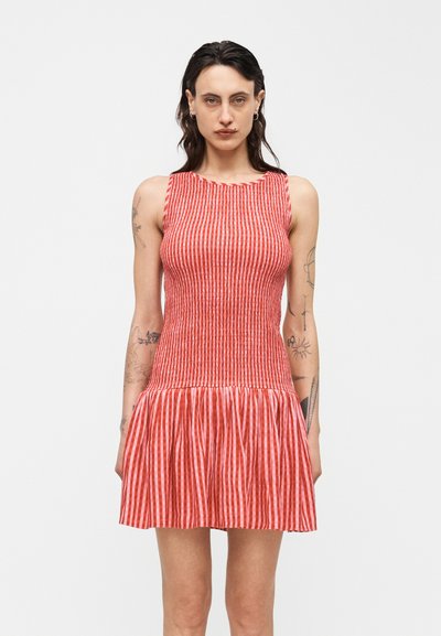 Mujer de cabello oscuro y tatuajes que lleva un vestido sin mangas de rayas verticales rojas y blancas con una falda acampanada.