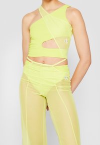 Nainen, jolla on yllään neonkeltaisen värinen yksioletuinen croptop, jossa on leikkuukohta, ja siihen sopivat läpinäkyvät korkeavyötäröiset housut, joiden alla on alushousut.