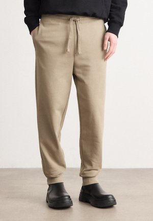 Mann trägt beige Jogginghose mit Kordelzug und Bündchen an den Knöcheln sowie schwarze klobige Slipper und steht auf einem glatten Boden vor einer schlichten Wand.