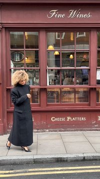 Lungo cappotto nero con design doppiopetto e ampio colletto, abbinato a scarpe nere con tacco alto. Esterno di un pub rosso con lettering dorato.