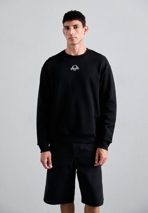Schwarzer Pullover aus weichem Stoff, mit einem gerippten Halsausschnitt und Bündchen. Enthält ein kleines Grafikdesign auf der Brust. Wird mit schwarzen Shorts getragen.