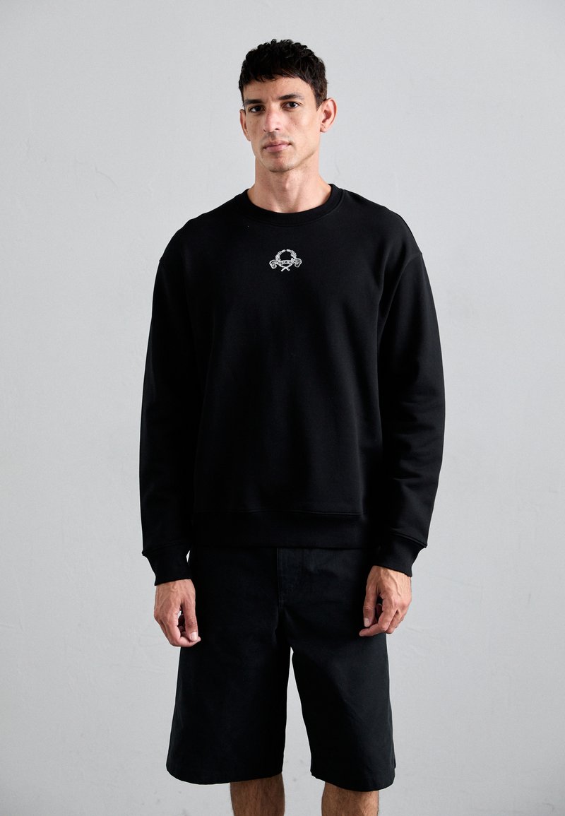 Svart sweatshirt i mjukt tyg, med en ribbad halsringning och mudd. Inkluderar en liten grafik på bröstet. Bärs med svarta shorts.