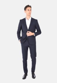 Completo blu con un design aderente, realizzato in tessuto morbido. Abbinato a una camicia bianca e scarpe in pelle nere. Presenta una chiusura a un bottone.