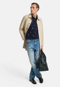 Cappotto beige su polo blu navy con motivo a veliero, jeans azzurri chiari, scarpe in pelle nere e borsa nera a tracolla, posa in piedi.