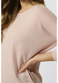 Femme aux longs cheveux portant un pull en maille rose clair, une main posée sur la hanche, sur un fond uni.
