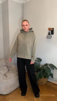 Světlé zelené mikinové tričko s kapsou na přední straně, kapucí se stahovací šňůrkou a dlouhými rukávy. Kombinováno s černými kalhotami s širokými nohavicemi a černou obuví.