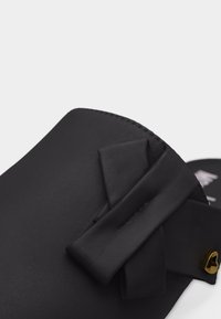 Μάσκα προσώπου από μαύρο ύφασμα με διπλωτή λεπτομέρεια φιόγκου και ένα μικρό χρυσό μενταγιόν σε σχήμα καρδιάς σε ουδέτερο φόντο.