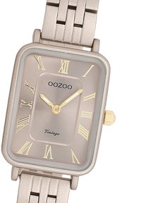 Montre rectangulaire en or rose avec un bracelet métallique, présentant un cadran gris clair, des chiffres romains en or et une couronne pour régler l'heure.