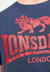 Sötétkék pamut póló, amely középen egy megkopott vörös oroszlán grafikát és vastag betűtípussal írt "Lonsdale London" feliratot tartalmaz.
