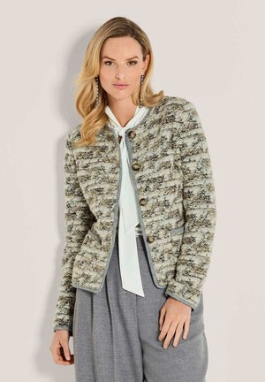 Frau trägt eine strukturierte, geknöpfte Strickjacke, eine weiße Bluse mit gebundenem Kragen, graue, plissierte Hosen und lange, hängende Ohrringe.