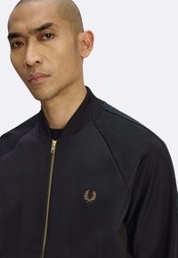 Giacca bomber nera con chiusura a zip e colletto a costine. Logo dorato sul petto a sinistra, tessuto con motivo sulle spalle e texture liscia.