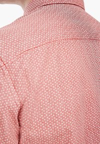 Camisa roja con un estampado de pequeños shapes geométricos blancos, cuellos estándar y una textura suave, visible parcialmente desde atrás.