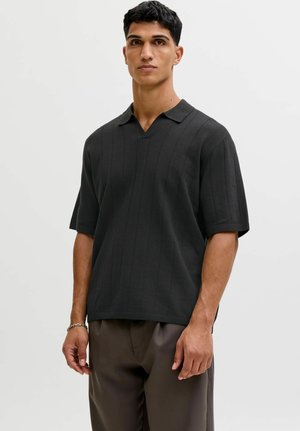 Mees seisab, kandes lühikeste varrukatega musta soonikkoega polo särki ja tumedad pruunid püksid, taust on lihtne.