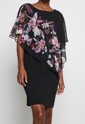 Robe noire avec superposition florale asymétrique transparente présentant des fleurs roses, blanches et violettes, portée par une personne debout sur un fond uni.