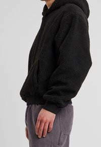 Felpa nera in pile con superficie testurizzata e soffice, dotata di cappuccio con coulisse e polsini a costine. Abbinata a pantaloni in velluto a coste grigio chiaro.