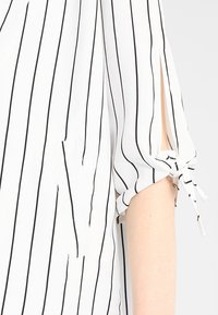 Blouse blanche à rayures noires verticales, manches courtes avec fentes latérales nouées en petits nœuds, et une poche verticale à l'avant.