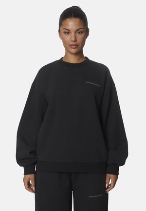 Donna con capelli scuri raccolti indossa una felpa nera oversize e pantaloni abbinati con una sottile scritta di branding.