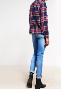 Camicia a quadri blu navy e rossa con bottoni, jeans aderenti blu e stivaletti neri alla caviglia. La camicia ha maniche lunghe e tasche sul petto.