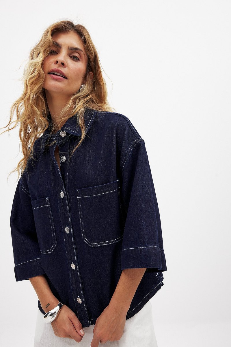 Kvinde med langt bølget blond hår iført en mørkeblå denimjakke med store forlommer og trekvartærmer mod en hvid baggrund.
