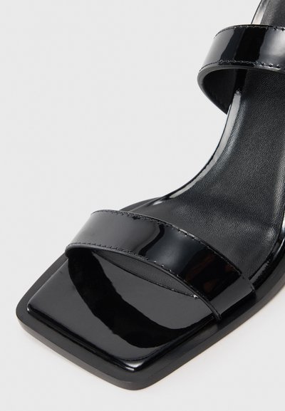 Sandal din piele lacuită neagră cu vârful pătrat, curea frontală și curea pentru gleznă, pe fundal alb, arătând detalii de aproape.