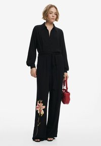 Combinaison noire à manches longues avec fermeture à boutons et ceinture à la taille, ornée d'un motif de broderie florale sur la jambe gauche. Accompagnée d'un sac à main rouge.