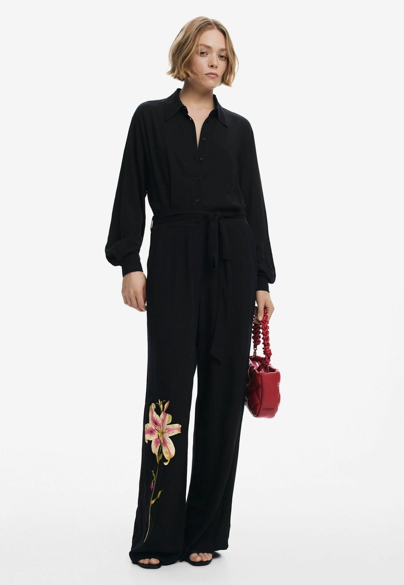 Combinaison noire à manches longues avec fermeture à boutons et ceinture à la taille, ornée d'un motif de broderie florale sur la jambe gauche. Accompagnée d'un sac à main rouge.