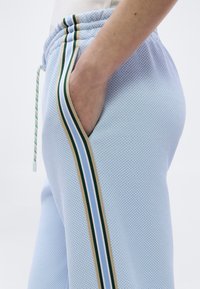 Χέρι ατόμου στην τσέπη φωτεινών γαλάζιων υφασμάτινων παντελονιών με λεπτομέρεια πλαϊνής ρίγας σε πράσινο, μπεζ και μπλε και ελαστικό κορδόνι στη μέση.