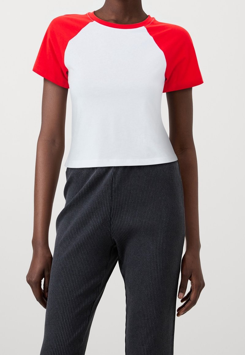 Personne portant un T-shirt blanc court avec des manches et un col rouges, associé à un pantalon côtelé foncé, visible du cou aux hanches.