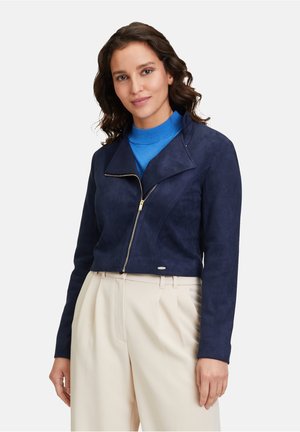 Marineblaues Wildlederjackett mit einem kurzen Design, breitem Kragen und einem Reißverschluss vorne; kombiniert mit einem blauen Rollkragenpullover und beigen Hosen.