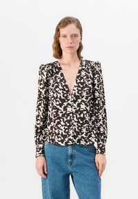Donna con capelli corti e mossi che indossa una blusa a maniche lunghe bianca e nera con bottoni e jeans blu a vita alta.