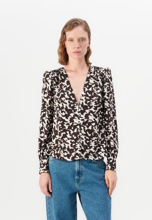 Woman with short wavy hair wearing a black and white patterned long-sleeve blouse with buttons and high-waisted blue jeans.