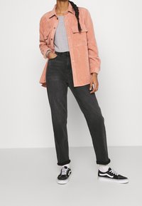 Korallfärgad kordflortsskjorta, grå t-shirt, högmidjade svarta jeans och svarta sneakers med vita sålar. Modell står mot en vit bakgrund.