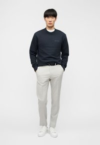 Νεαρός άνδρας που στέκεται με τα χέρια στις τσέπες, φοράει μαύρο φούτερ, ανοιχτόγκρι παντελόνι, μαύρη ζώνη και λευκά αθλητικά παπούτσια.