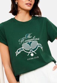 Zaļš kokvilnas T-krekls ar balto grafiku, kurā attēloti krusteniski tenisa raketi un teksts "The Hamptons Social Club", apaļš kakla izgriezums un īsas piedurknes.