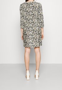 Blumenkleid mit dunklem Hintergrund, Dreiviertelärmeln, gerafftem Taillenbereich und knielanger Länge. Kombiniert mit weißen Blockabsatzsandalen.