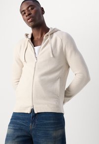 Lyse beige zip-up hettegenser med hette, ribbet tekstur og sidelommer. Glidelås foran og justerbare snorer i hetten.