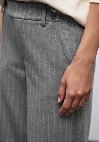 Pantaloni grigi a righe sottili con una texture liscia, dotati di un frontale piatto, due bottoni e una tasca laterale. Mano appoggiata sul tessuto.