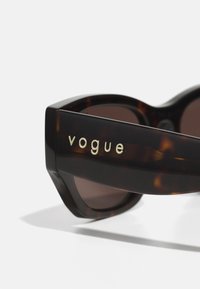 Rudos tortų šešėliai saulės akiniai su per dideliais geometriniais rėmeliais. Šakos turi išrašytą "vogue" auksu. Lęšiai yra tamsiai rudi.