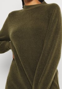 Maglione verde oliva con una finitura morbida e testurizzata, caratterizzato da un collo alto e maniche lunghe. La vestibilità è rilassata, con dettagli di cucitura minimi.
