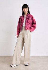 Giacca in denim rosa sbiadito con due tasche sul petto, abbinata a pantaloni beige larghi e scarpe da ginnastica bianche, in posa su uno sfondo neutro.