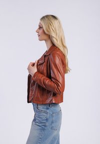 Bruine leren motorjack met een getailleerd ontwerp, reverskraag en ritssluiting aan de voorkant. Gecombineerd met lichtgewassen denim jeans.