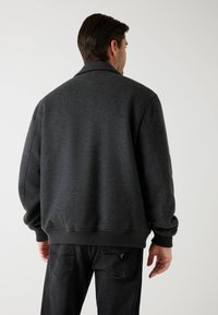Jachetă gri dintr-un amestec de lână, cu un guler înalt, manșete și tiv ribbate. Spatele prezintă o croială lejeră cu o suprafață netedă și texturată.
