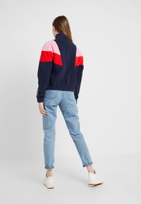 Námořnická fleecová bunda s barevně rozdělenými panely v růžové, červené a námořnické modré barvě, vysokým límcem a elastickým pasem. V kombinaci s světle modrými džíny a bílými teniskami.