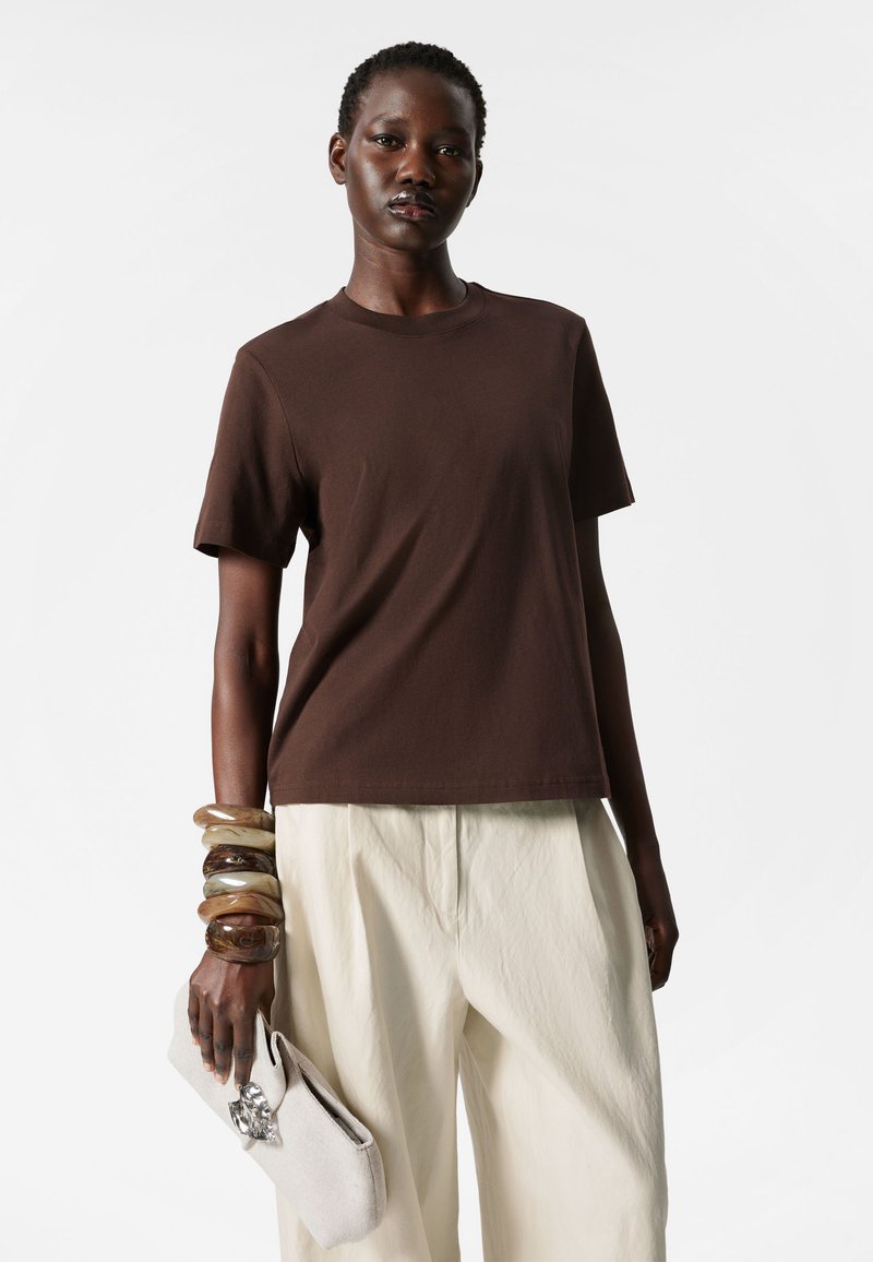 Brown cotton t-shirt with short sleeves, paired with cream-colored loose trousers. Accessories include multiple wooden bangles and a light clutch.